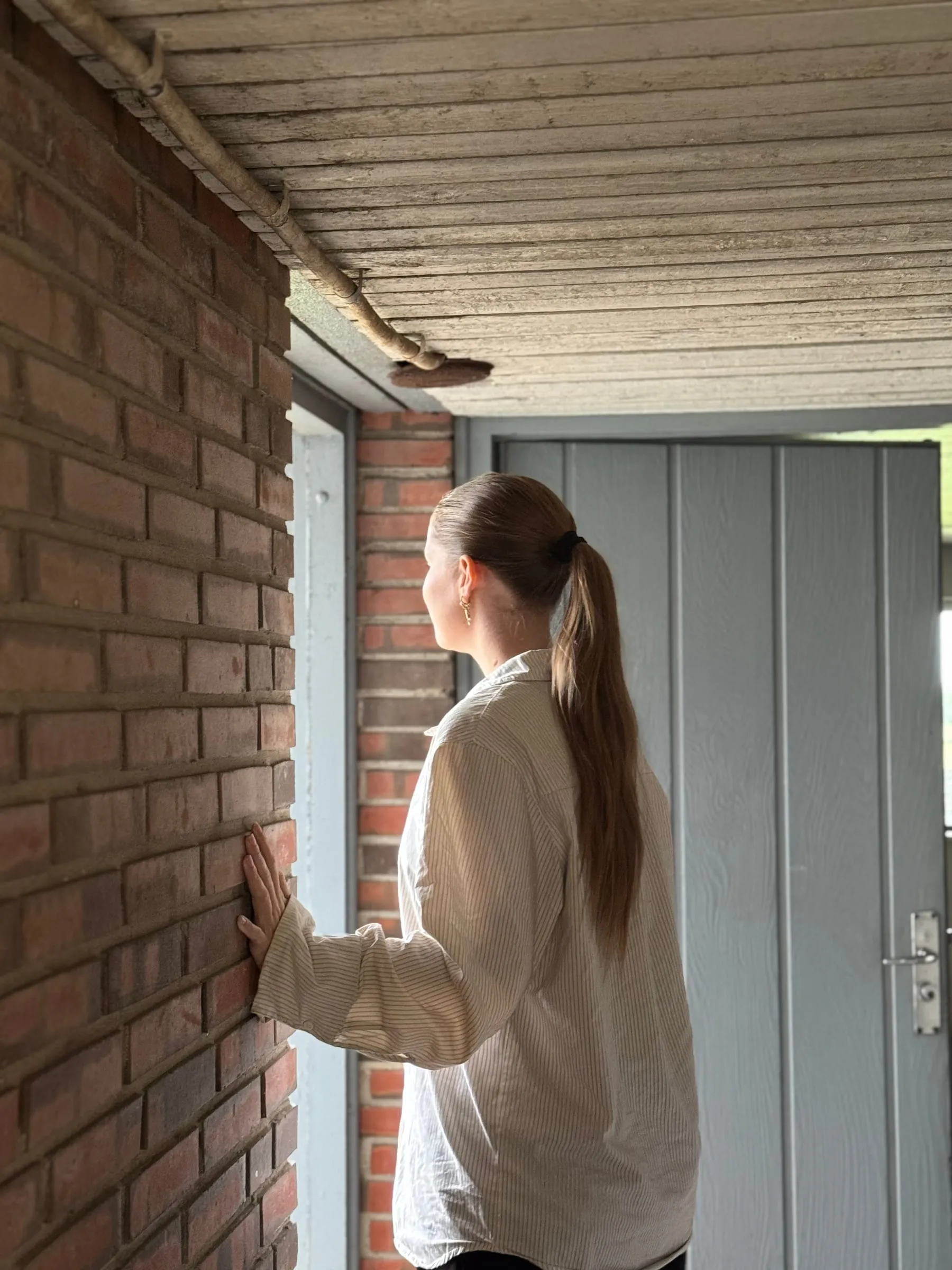 Sarah Tarp med hånden på husets murstensfacade, kiggende mod hoveddøren